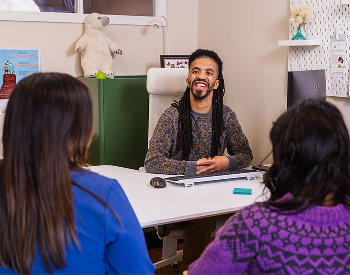 A cheerful professional with long hair and a beard sits at a white desk, engaging in a lively conversation with two individuals seated across from him. A playful stuffed polar bear is placed on top of a green cabinet in the background.