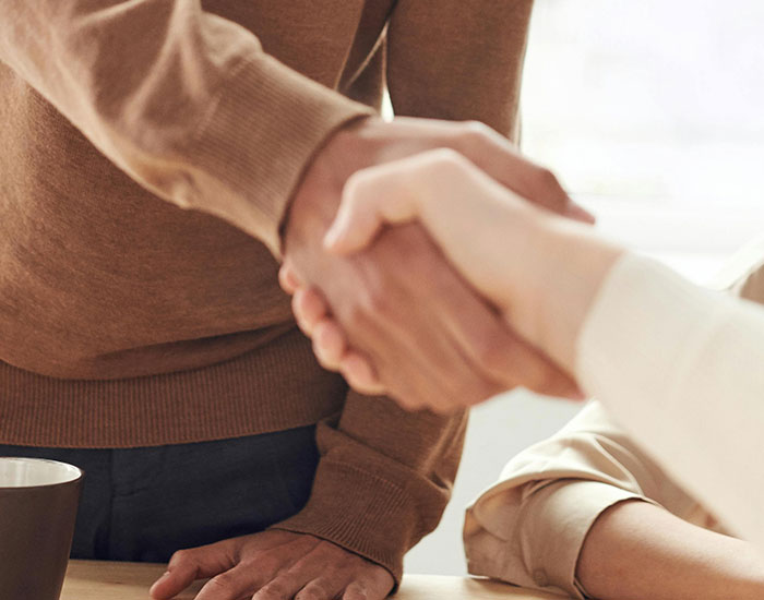 A close up of two people in neutral coloured clothing shaking hands. They are around a table with a coffee cup on it.