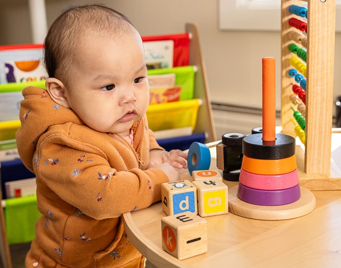 A baby wearing an orange hoodie sits at a table with various toys. The child plays with wooden blocks and a stacking ring toy. Colorful books and an abacus are visible in the background.