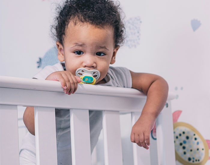 A young child stands in a crib, holding onto the railing with one hand. The child has curly hair and is wearing a white outfit, while using a pacifier. The background features colorful, playful decorations.