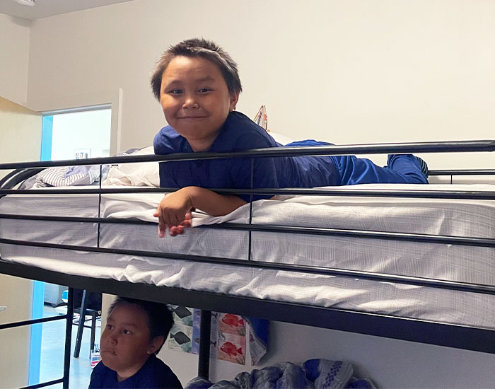 Two children are interacting in a bedroom with bunk beds. The child on the top bunk, dressed in a blue shirt, is smiling at the camera. The lower bunk has another child who is looking up at the top bunk. The room has a door to the left and various items on the beds.