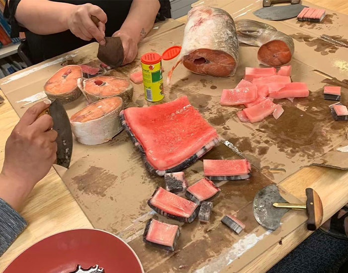 Two people are cutting large pieces of frozen fish on a table. One person is using a large knife, and the other is holding a cleaver. Various cuts of fish are spread out, alongside some knives and a can of seasoning.