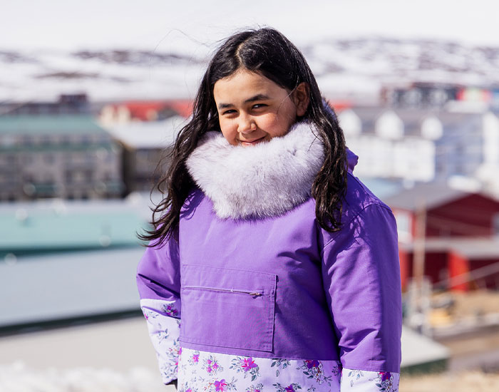 A young person with long, dark hair smiles while wearing a purple winter coat with a light fur collar and floral accents. They are standing outdoors with snowy buildings in the blurred background.