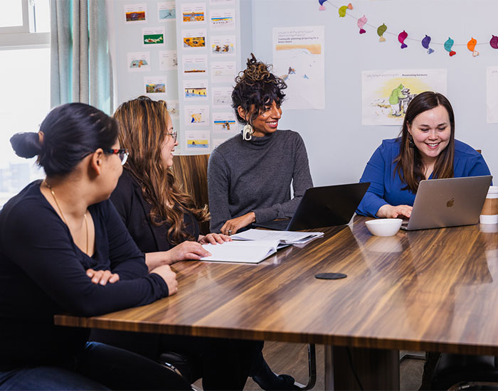 Four people are sitting around a wooden table in a well-lit room, collaborating. Two have laptops, one is holding papers, and another has a notebook. The background features posters on the wall and colourful decorations. Everyone appears engaged in discussion.