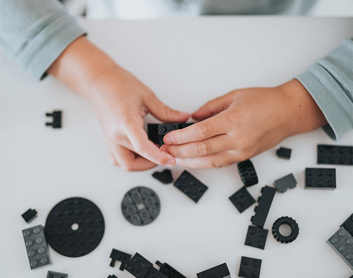 A person is assembling various black plastic building blocks on a white surface. Their hands are in the process of connecting pieces, with multiple blocks scattered around. The person is wearing a light blue long-sleeved shirt.