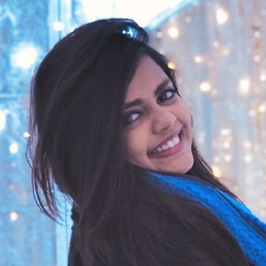 A young person with long, dark hair smiles warmly at the camera. They are wearing a dark top and a blue scarf. The background features soft, glowing lights, creating a festive and cozy atmosphere.