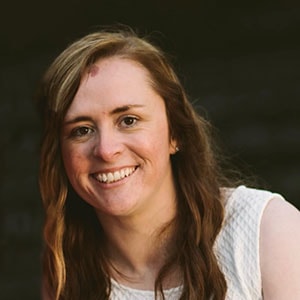 A woman with long, wavy brown hair smiles at the camera. She is wearing a white sleeveless top and there is a blurred dark background. She has a distinctive mark on her forehead.