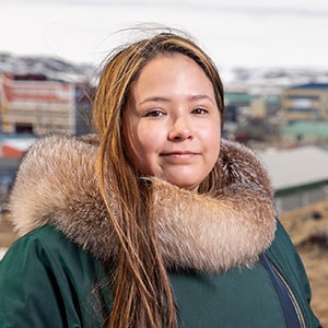 A woman with long brown hair wearing a green coat with a fur collar is looking at the camera. The background shows a town with multicolored buildings and snow-covered hills in the distance.