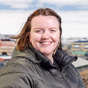 A woman with shoulder-length hair is smiling at the camera. She is wearing a dark green puffer jacket and standing outdoors with buildings in the background under a partly cloudy sky.