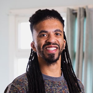 A person with long dark dreadlocks and a beard is smiling at the camera. They are wearing a patterned sweater, and there is a window with curtains in the background.