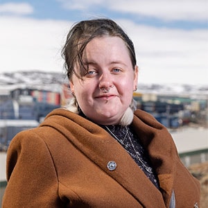 A person with short hair and visible piercings is outdoors wearing a brown coat and a black patterned shirt. The background shows a scenic view with buildings and snow-covered hills under a partly cloudy sky.