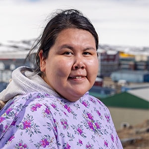 A woman with long dark hair smiles at the camera. She is wearing a light purple floral top and a gray hooded jacket underneath. The background shows a blurred view of buildings and snow-covered structures, suggesting a cold, urban environment.