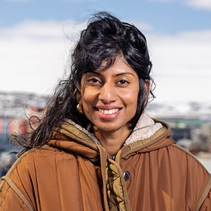 A woman with dark, wavy hair and wearing a brown winter coat smiles at the camera. The background is slightly blurred, showing a hint of snowy terrain and possibly some buildings.