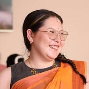 A person with long braided hair smiles warmly, wearing glasses and a bindi on their forehead. They are dressed in a traditional outfit, including an orange saree and gold jewelry, set against a softly blurred background.