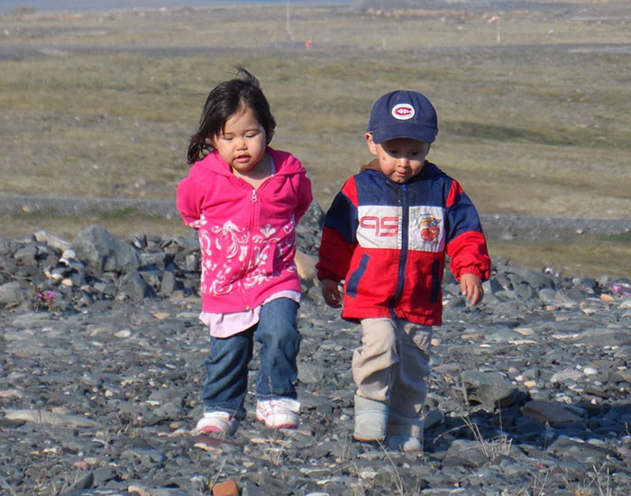 Two young children walk together on a rocky terrain. The girl on the left is wearing a pink jacket, while the boy on the right sports a red and blue jacket with a baseball cap. The background shows a vast, open landscape with a clear sky.